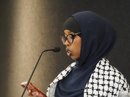 Woman gives reads prayer from phone at pulpit for close of Ramadan at Interfaith Iftar